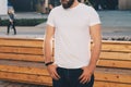 Front view. Young bearded hipster man dressed in white t-shirt and sunglasses is stands on city street. Mock up. Royalty Free Stock Photo