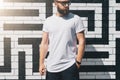 Front view. Young bearded millennial man dressed in white t-shirt and sunglasses is stands against brick wall. Mock up.