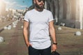 Front view. Young bearded millennial man dressed in white t-shirt and sunglasses is stands on city street. Mock up. Royalty Free Stock Photo