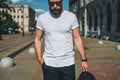 Front view. Young bearded millennial man dressed in white t-shirt and sunglasses is stands on city street. Mock up. Royalty Free Stock Photo