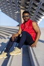 Front view of a young black man wearing sunglasses sitting on staircase in a sunny day while looking camera Royalty Free Stock Photo