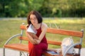 Front View of Young Girl in Eyesglasses and Long Red Dress Sitting on the Bench in the City Park and Reading Some Book. Royalty Free Stock Photo