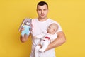 Front view of young handsome father wearing white casual t shirt standing isolated over yellow background, holding newborn baby Royalty Free Stock Photo