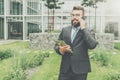 Front view. Young happy businessman in suit and tie is standing outdoor,holding tablet computer and talking on his cell Royalty Free Stock Photo