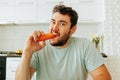 Front view of a young man biting a raw carrot with a ferocious and unhappy expression.