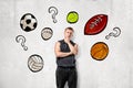 Front view of young man in sleeveless hoody standing with hand rubbing chin at wall with drawings of different sport