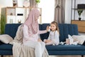 Front view of young pretty mother, Muslim woman in hijab, sitting on blue couch at home and wiping face of her cute