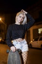 Front view of young pretty woman looking camera in shorts while standing on the street holding a skateboard at night in the city Royalty Free Stock Photo