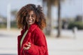 Front view of a young smiling curly afro woman standing outdoors while smiling and listening music by earphones in a sunny day Royalty Free Stock Photo