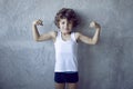 Frontal of a little curly haired red boy in holding fists up and showing biceps on concrete background Royalty Free Stock Photo