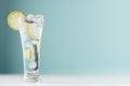 Frosty transparent lemonade with lemon slices, ice cubes and mineral water in elegant glass on white wood table, mint color wall.