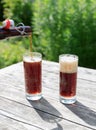 Frothy dark beer pouring into tall glasses from a brown glass bottle in summer garden on rustic wooden table