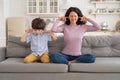 Frowning mom and son sit on couch with closed eyes and cover ears from noisy music or fight sounds