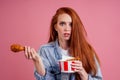 Frowning vegetarian redhair ginger woman holding fried chicken`s leg and a bucket of fast food french fries potatoes and Royalty Free Stock Photo