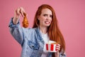 Frowning vegetarian redhair ginger woman holding fried chicken`s leg and a bucket of fast food french fries potatoes and Royalty Free Stock Photo