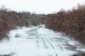 A frozen river covered with snow with lots of leafless trees on the sides. Ice is melting. An early spring