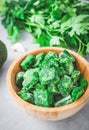 Frozen spinach cubes in a wooden bowl with blurred fresh parsley and a clove of garlic on a gray .