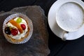 Fruit cake with cherries, grapes and plum stands on a wild stone next to a white cup of coffee with a foam Royalty Free Stock Photo