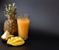 Fruit juice in a tall glass on a black background, next to pieces of ripe pineapple and mango Royalty Free Stock Photo