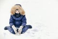 A frustrated child with sad eyes in a jumpsuit with a fur hood and ruddy cheeks from frost. Sits on snow a winter