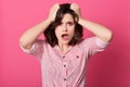 Frustrated confused Caucasian woman keeping hands in her hair, posing with widely opened mouth, wearing striped shirt, being in Royalty Free Stock Photo