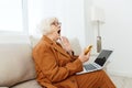 a frustrated elderly woman is sitting in an apartment on a beige sofa in a brown pantsuit and holding a laptop on her Royalty Free Stock Photo