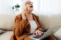 a frustrated elderly woman is sitting in an apartment on a beige sofa in a brown pantsuit and holding a laptop on her Royalty Free Stock Photo