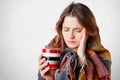 Frustrated ill young woman covered in warm blanket, keeps hand on temple, suffers from terrible headache, drinks hot beverage, tri Royalty Free Stock Photo