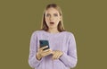 Frustrated shocked young blondy girl in purple sweater with smartphone in hands on khaki background.