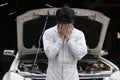 Frustrated stressed young mechanic man in white uniform covering his face with hands against car in open hood at the repair garage Royalty Free Stock Photo