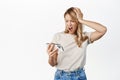 Frustrated and upset blond girl looking at smartphone, trouble with mobile phone, watching upsetting news on cellphone