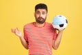 Frustrated young bearded man in red t-shirt holding in hand soccer ball in medical mask, problems with the football championship, Royalty Free Stock Photo