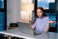 Frustrated young woman confused by laptop computer problem sitting at desk in home office near window evening at late