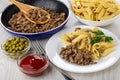 Frying pan with fried mince, bowl with pasta, green peas, ketchup, plate with macaroni, fried mincemeat, fork on table