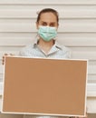 A young woman in a mask holds an empty cork board in her hands, standing on a white background. Copy space on whiteboard for image Royalty Free Stock Photo