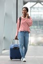 Full body happy young black woman walking with suitcase in airport terminal with cellphone Royalty Free Stock Photo