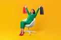 Full body length shot of contented shopaholic woman sitting on chair and raising hands with shopping bags, free space