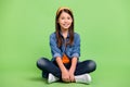 Full body photo of cute small girl sit wear shirt jeans hairband sneakers isolated on green background Royalty Free Stock Photo