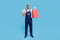 Full body portrait of excited happy courier in uniform holding shopping bags and credit card, studio shot