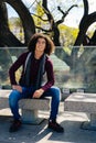 Full body portrait of a smiling young curly haired Latino man sitting on a concrete bench in the shade of an old tree. Vertical Royalty Free Stock Photo