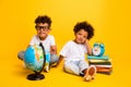 Full body portrait of two clever small funny kids sit floor education supply globe book clock isolated on yellow color