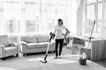 Full body portrait of young woman in white shirt and jeans cleaning carpet with vacuum cleaner in living room, copy space. Royalty Free Stock Photo