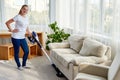 Full body portrait of young woman in white shirt and jeans cleaning carpet with vacuum cleaner in living room, copy space. Royalty Free Stock Photo