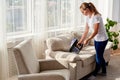 Full body portrait of young woman in white shirt and jeans cleaning sofa with vacuum cleaner in living room, copy space. Housework Royalty Free Stock Photo