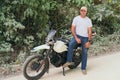 Full body of serious young ethnic biker in casual clothes and cap standing on motorcycle