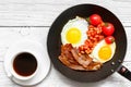 Full english breakfast in frying pan with fried eggs, bacon, beans, tomatoes and coffee on white wooden background Royalty Free Stock Photo