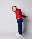 Full growth portrait of frolic blond kid boy in red and blue sport suit standing sideways to us with arms outstretched Royalty Free Stock Photo