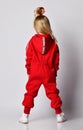 Blonde kid girl in red jumpsuit with hood and cool hairstyle stands with her hands in pockets. Back view. Full-growth.