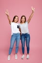 Full lengh image of charming two young twin sisters with beautiful smile with hands up showing peace gesture over pink Royalty Free Stock Photo