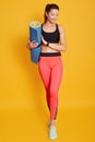 Full lengh shot of attractive young woman with yoga mat, stands against yellow studio background with some copy space, looks down